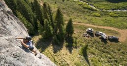 Photo of man climbing rocks with two pickup trucks and travel trailers below.