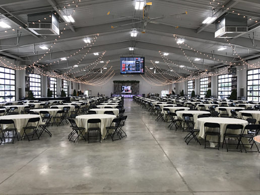 A photograph of Interior of the Peter B. Orthwein Pavilion at the RV/MH Hall of Fame in Elkhart, Indiana. There are 40+ round tables lined up in neat rows. The tables have folding chairs arranged around them. There are strings of white lights decorating the ceiling of the hall. A stage is located across the room from where the camera is set up.