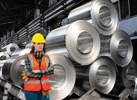 A picture of a woman in construction gear standing in front of about a dozen big rolls of sheet metal stacked up in several piles