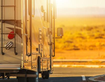 A picture of an RV driving down the road away from the camera with the setting sun glaring off the side of the RV