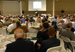 A photograph from the audience's perspective of a man standing at a podium addressing a conference hall full of people. This is the Recreational Vehicle and Motorhome Hall of Fame Induction Dinner