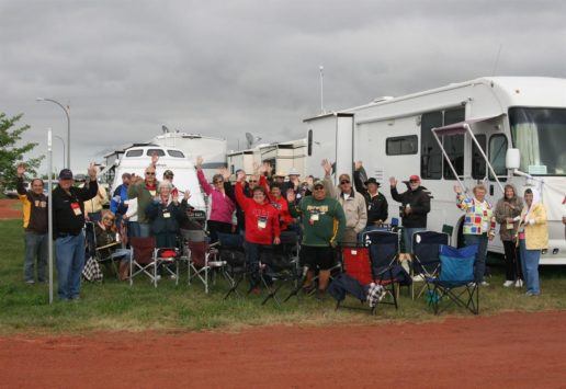 A photograph of about 24 people standing and sitting in lawn chairs in front of their RVs on a cloudy day.