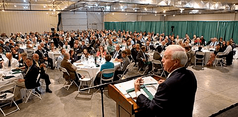 A photograph from the stage's perspective of a man standing at a podium addressing a conference hall full of people. This is the Recreational Vehicle and Motorhome Hall of Fame Induction Dinner
