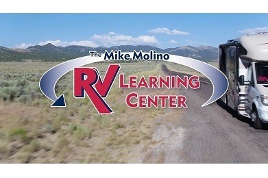 A picture of a Type C RV driving down a highway with prairie brush on the side of the road and mountains in the background. A graphic saying, "The Mike Molino RV Learning Center" has been superimposed on the image.