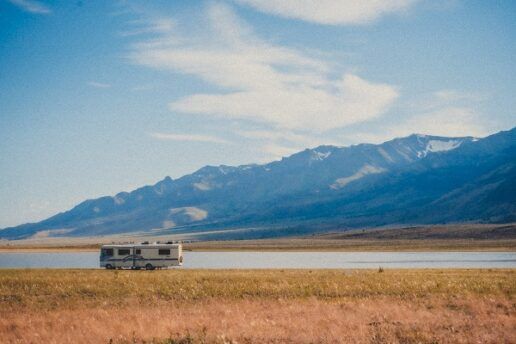 A picture of a lone motorhome parked next to a lake. Yelllow grass is in the foreground, and a clear sky over blue mountains are in the background behind the lake.