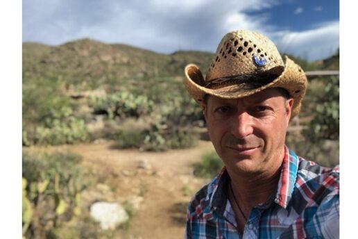 A photograph of Scott Watson of Go Small Live Large. Watson is wearing a straw cowboy hat and a plaid shirt. The desert opens up behind him. One big cloud is overtaking the sky.