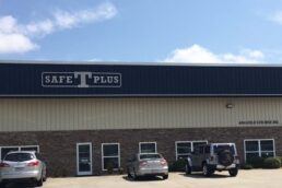 A photograph of the front of a building that says "Safe T Plus" in bold lettering on the side. Three vehicles are parked in front of the building, and the sky behind the building is blue with puffy clouds.