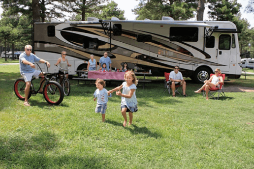 A photo of a family spending time together in the grass outside of a type A motorhome. A couple of adults are sitting on bikes. Two children are running on the grass. Four people are sitting at a picnic table, and two more people are lounging in camping chairs off to the side.