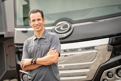 A picture of race car driver Helio Castroneves, wearing a gray American Coach polo shirt, in front of an American Coach motorhome