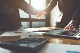 A picture of two people in suits shaking hands as they are standing up from a conference table.