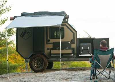 A picture of Alcom's Overland Camper set up on an off-road site with a camper in a chair in front of it.