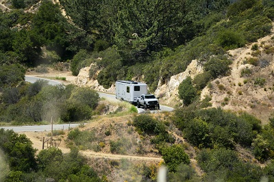 A picture of the 2023 Living Vehicle trailer being towed by a truck on a road through the hills