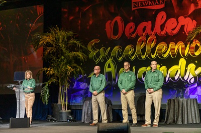 A picture of three men in green shirts and khakis and a woman at a podium, all are onstage in front of a backdrrop that reads Dealer Excellence Awards