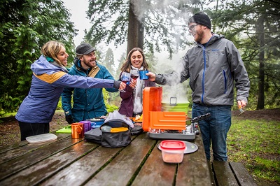 A stock photo of four adult campers at a table using GSI Outdoors equipment while surrounded by large pine trees.