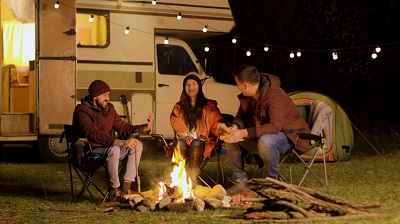 An RVIA stock image of three campers at night, sitting in camping chairs around a fire and laughing, with their Type C motorhome behind them