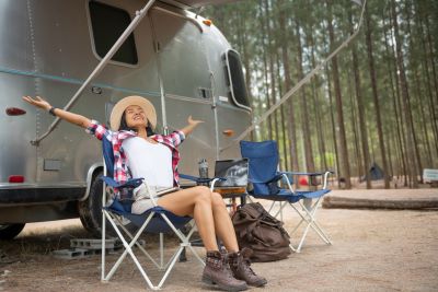 A picture of a woman sitting outside an Airstream-type travel trailer in a camp chair with her arms thrown wide in exhuberance