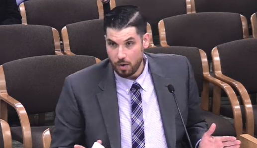 A picture of RVIA's Nick Rudowich in a suit in a legislative setting with rows of seats behind him