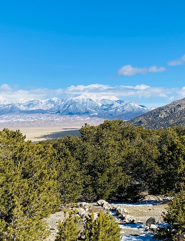 A picture of the snow-capped Great Sand Dunes of Colorado from a distant trailhead with a blue sky and snow covered peaks in the background.