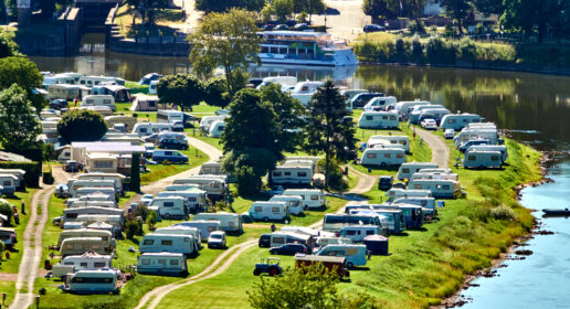 A picture of a crowded campground showing an arial view of RVs lined up in rows on verdant ground next to a winding river.