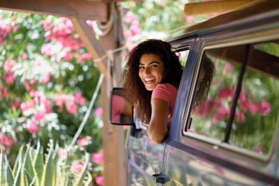 A picture of a woman in a pink shirt leaning out the window of a Type B camper van with spring flowers in the background