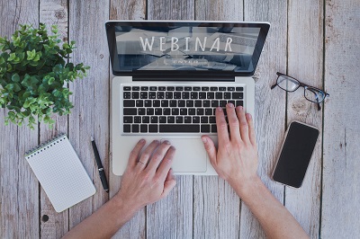 An overhead picture of a person working on a laptop with a notepad, plant, phone and pen on their desk and the word Webinar on the screen