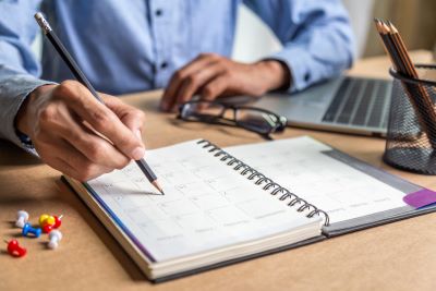 A picture of a male figure in a crisp blue shirt penciling notes into a paper day planner with his laptop at his left elbow