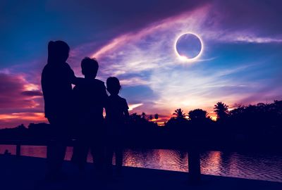 A picture of a total solar eclipse with a family silhouetted in the half-dark in front of a lake.