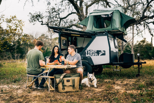 A picture of the Taxa Tiger Moth travel trailer in the background with three young adults playing cards in the foreground, their white dog happily laxing on the ground at their sides