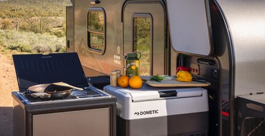 A picture of a 2023 InTech Flyer adventure trailer slide out kitchen showing a grill on the far left with a pan on it and a Dometic electric cooler with fruit, vegetables and a cutting board on it.