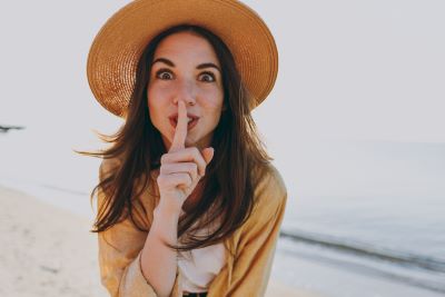 A picture of a young woman in a hat on a beach holding her finger to her lips in a universal symbol for "shhh" or hush
