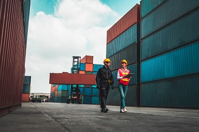 A picture of two people walking through a shipping yard piled high with containers. One person has a clipboard