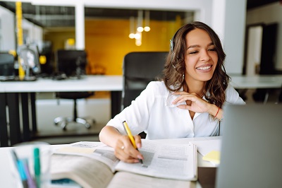 A picture of a young woman wearing earphones joining in on a webinar and taking notes.
