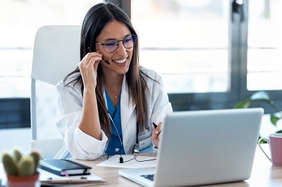 A picture of a young Asian woman in a doctor's coat talking on a headset to a patient via her laptop symbolizing telehealth