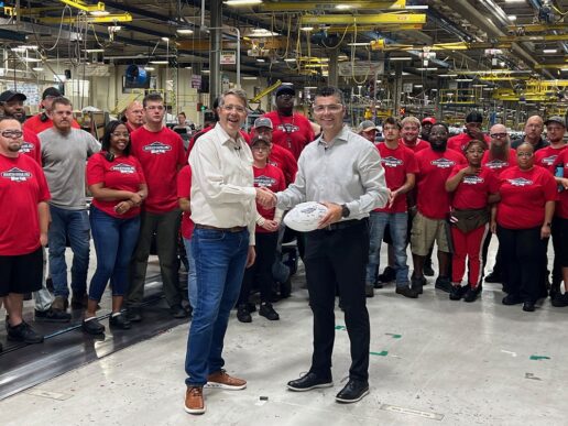 A picture of (L to R) Freightliner Custom Chassis Corp. President and CEO Jeff Sather and Tiffin Motorhomes President Leigh Tiffin holding a signed football in front of FCCC employees wearing red work shirts.
