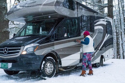 A stock image of a woman outside a Type C motorhome parked in the snow while snow falls.