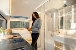 A picture of a woman standing in the galley of a Pebble Flow electric travel trailer. Behind her is a transparent wall to the bathroom and the rear bedroom/office.