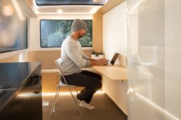 A picture of a man sitting at the desk in the rear office of the Pebble Flow travel trailer. The desk is attached to the underside of the room's Murphy bed.