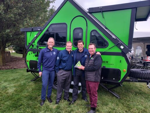 A picture of Blue Compass RV CEO Jon Ferrando receiving an award from Aliner executives in front of an Aliner A-frame RV.