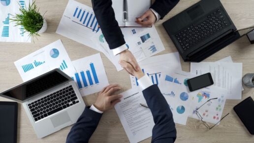 A stock photo of two men shaking hands over a table covered in charts, laptops, infographics and graphs.